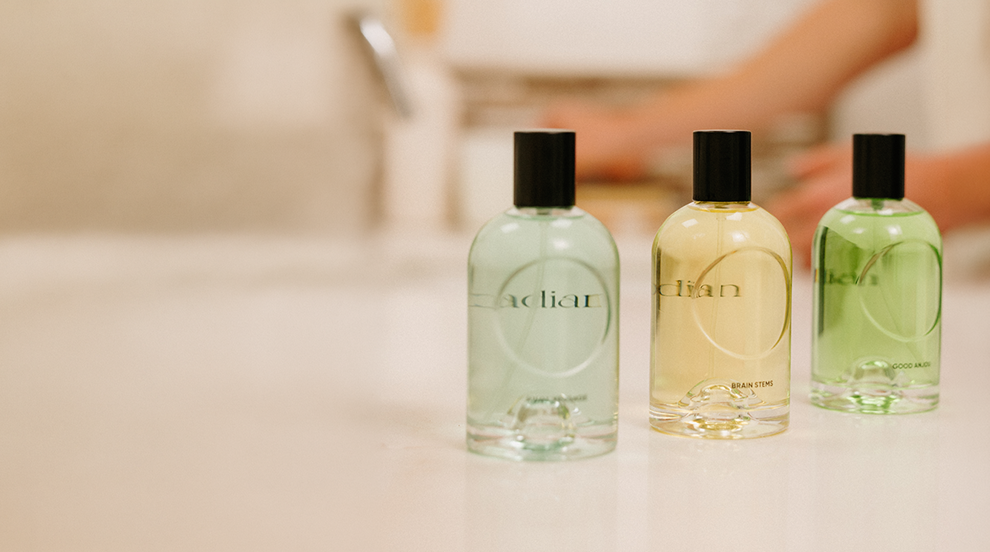 Three clear glass bottles of Cirqadian fragrance with black caps lined up on a white countertop, with a blurred bathroom background showing a person's hands and arm.
