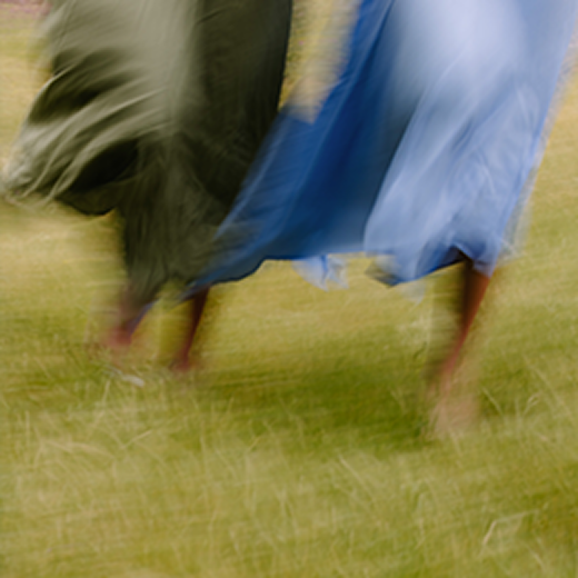 A motion-blurred outdoor scene showing two people barefoot on grass, one wearing a dark green garment and the other wearing a flowing light blue garment.